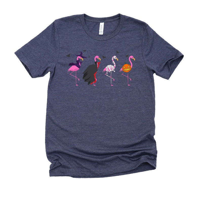 May include: A navy blue t-shirt with a graphic of five pink flamingos dressed in Halloween costumes. The flamingos are wearing a witch hat, a vampire cape, a mummy costume, a skeleton costume, and a pumpkin costume.