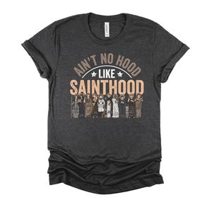 Pu&ograve; includere: T-shirt grigio scuro con la scritta "Ain't No Hood Like Sainthood" in un design circolare sopra una fila di figure illustrate. La maglietta &egrave; realizzata in materiale morbido e ha le maniche arrotolate.