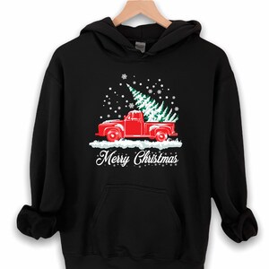 May include: Black hooded sweatshirt with a red pickup truck carrying a Christmas tree. The truck is driving through a snowy scene with snowflakes falling. The text "Merry Christmas" is printed below the truck.