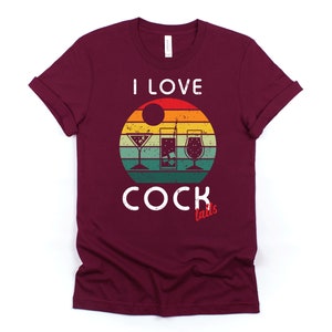 Op de afbeelding: Een maroon t-shirt met een vintage-stijl grafische afbeelding van een zonsondergang en drie cocktailglazen. De tekst "Ik hou van cocktails" is in wit op het shirt gedrukt.