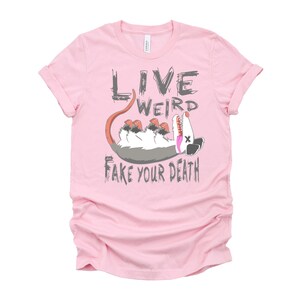 Op de afbeelding: Roze T-shirt met een afbeelding van een opossum die dood speelt en de tekst "Live Weird Fake Your Death."