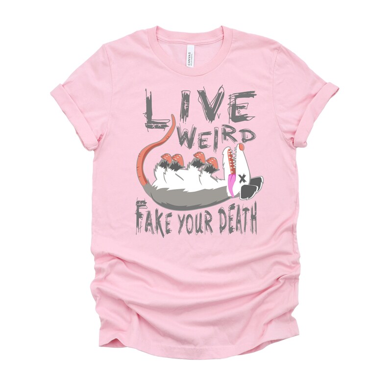 Op de afbeelding: Roze T-shirt met een afbeelding van een opossum die dood speelt en de tekst "Live Weird Fake Your Death."