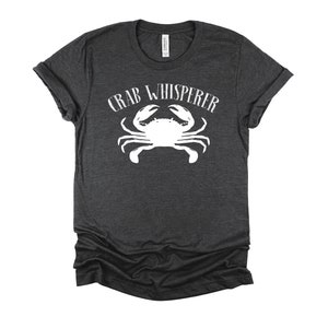 May include: A dark heather grey t-shirt with a white crab graphic and the text "CRAB WHISPERER" printed on the front.