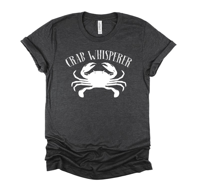 May include: A dark heather grey t-shirt with a white crab graphic and the text "CRAB WHISPERER" printed on the front.