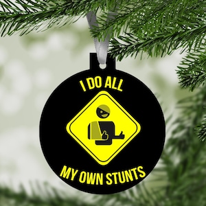 May include: Black round Christmas ornament with the text "I DO ALL MY OWN STUNTS". The ornament features a yellow diamond-shaped graphic of a person with an injured arm and a thumbs-up gesture.