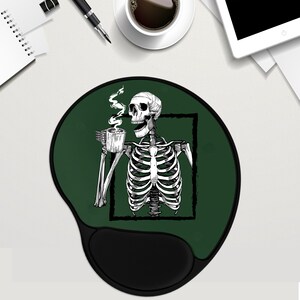 May include: Green round mouse mat with a black skeleton holding a cup of coffee. The skeleton is inside a white square frame. The mouse mat has a black wrist rest.