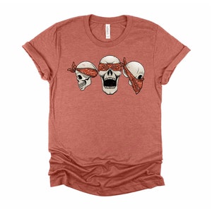 May include: A red heather t-shirt with a graphic of three skulls wearing bandanas. The middle skull is open-mouthed and the skulls on either side have their eyes covered by the bandanas.