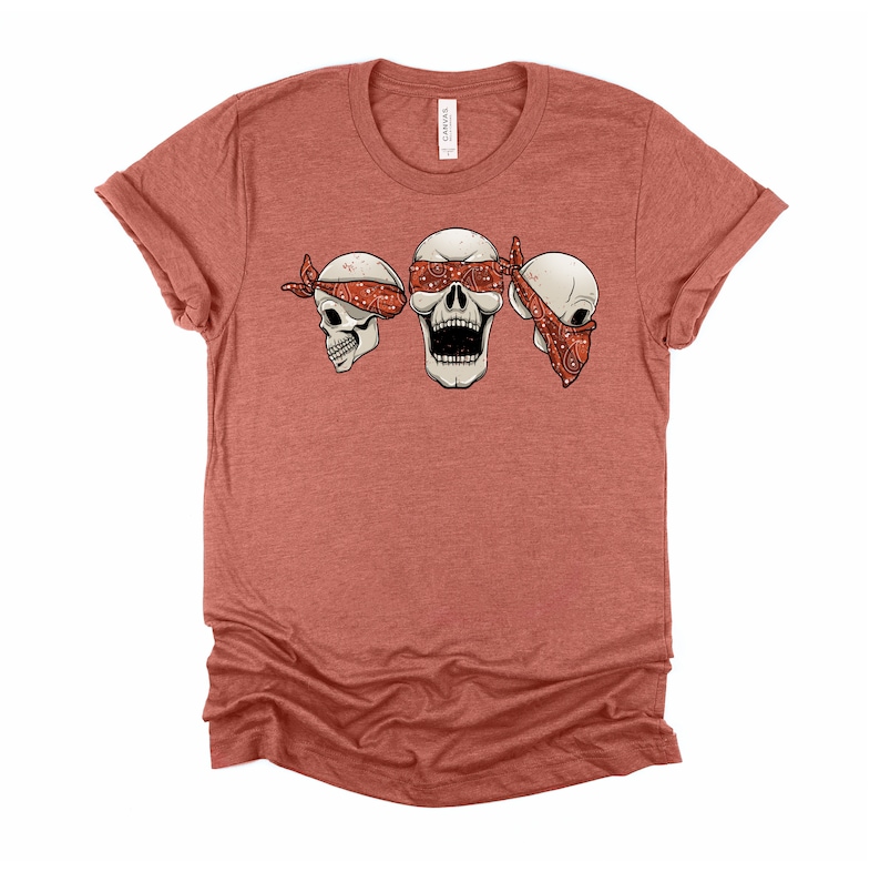 May include: A red heather t-shirt with a graphic of three skulls wearing bandanas. The middle skull is open-mouthed and the skulls on either side have their eyes covered by the bandanas.