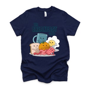 May include: A navy blue t-shirt featuring a graphic of breakfast foods, including a mug, orange, toast, egg, and bacon. The text "The Breakfast Club" is printed on the shirt.