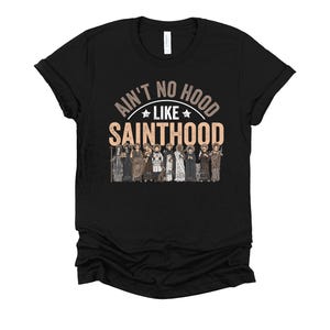 Pu&ograve; includere: T-shirt nero con la scritta "Ain't No Hood Like Sainthood" in un design circolare. Sotto il testo, un'illustrazione di figure in abiti religiosi. La maglietta &egrave; realizzata in materiale morbido.