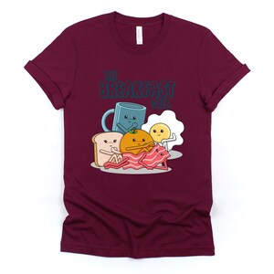 May include: A maroon t-shirt featuring a graphic of breakfast foods with faces. The text "The Breakfast Club" is printed on the shirt.