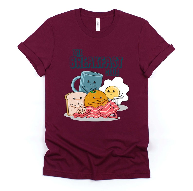 May include: A maroon t-shirt featuring a graphic of breakfast foods with faces. The text "The Breakfast Club" is printed on the shirt.