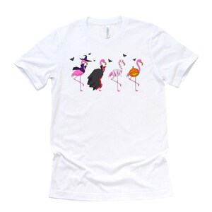 May include: White t-shirt with a graphic of four pink flamingos dressed in Halloween costumes. The flamingos are dressed as a witch, a vampire, a mummy, and a pumpkin.