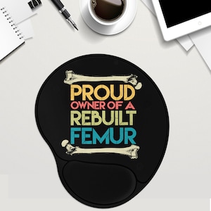 May include: Black ergonomic mouse pad with a wrist rest. The pad features the text "PROUD OWNER OF A REBUILT FEMUR" in a retro color scheme, with bone illustrations. The pad is on a white surface with a coffee cup, pen, and notepad.