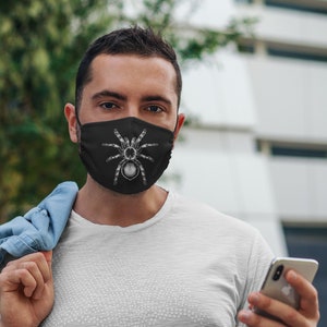 K&ouml;nnte beinhalten: Schwarze Gesichtsmaske mit einer detaillierten wei&szlig;en Illustration einer Vogelspinne. Die Maske wird von einer Person in einem wei&szlig;en T-Shirt getragen. Der Hintergrund ist verschwommen.