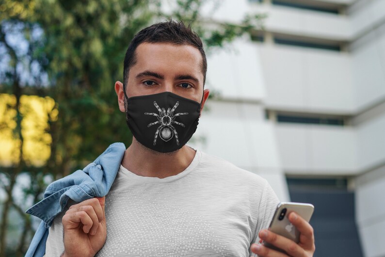 K&ouml;nnte beinhalten: Schwarze Gesichtsmaske mit einer detaillierten wei&szlig;en Illustration einer Vogelspinne. Die Maske wird von einer Person in einem wei&szlig;en T-Shirt getragen. Der Hintergrund ist verschwommen.