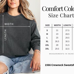 Op de afbeelding: Een donkergrijs Comfort Colors crewneck sweatshirt. De afbeelding bevat een maattabel met afmetingen in inches voor maten S t/m 3XL. De tekst "1566 Crewneck Sweatshirt" is ook zichtbaar.