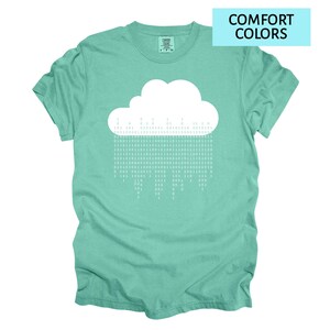Pode incluir: Camiseta verde-&aacute;gua com um desenho de nuvem branca e chuva de c&oacute;digo bin&aacute;rio. O texto "COMFORT COLORS" est&aacute; em um ret&acirc;ngulo azul claro. A camiseta tem gola redonda e mangas curtas. O tecido &eacute; macio.