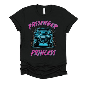 May include: Black t-shirt with a graphic of a blue and black off-road vehicle. The text "PASSENGER PRINCESS" is in pink and blue above and below the vehicle graphic. The shirt is made of a soft material.
