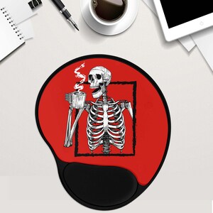 May include: Red mouse mat with a black skeleton holding a cup of coffee. The skeleton is inside a black square frame. The mouse mat has a black, rounded wrist rest.