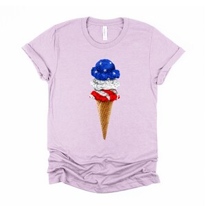 May include: A light purple t-shirt with a graphic of a three-scoop ice cream cone. The ice cream is decorated with red, white, and blue to resemble the American flag.