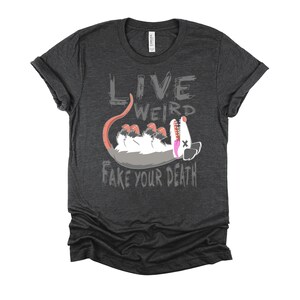 Op de afbeelding: Een donker grijs t-shirt met een afbeelding van een opossum die dood speelt en de tekst "Live Weird Fake Your Death".