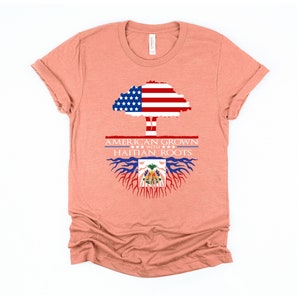 May include: A peach-colored t-shirt with a graphic design of a tree with the American flag as the leaves and Haitian flag as the roots. The text "AMERICAN GROWN WITH HAITIAN ROOTS" is printed on the shirt.