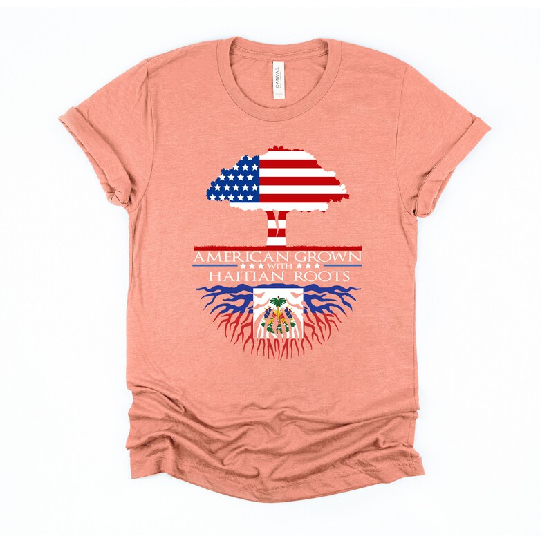 May include: A peach-colored t-shirt with a graphic design of a tree with the American flag as the leaves and Haitian flag as the roots. The text "AMERICAN GROWN WITH HAITIAN ROOTS" is printed on the shirt.