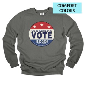 May include: A dark gray sweatshirt with a circular graphic that reads "WOMEN'S VOTE 1920-2020 100 YEARS 19TH AMENDMENT". The graphic is red, white, and blue with three white stars at the top. The words "COMFORT COLORS" are in a light blue rectangle.