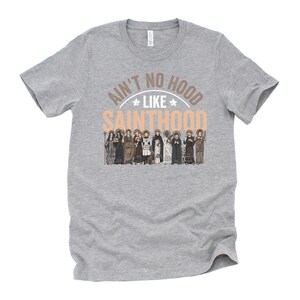 Pu&ograve; includere: T-shirt grigia con la scritta "Ain't No Hood Like Sainthood" in marrone chiaro e bianco. Il design include un'illustrazione di diversi santi in varie vesti.