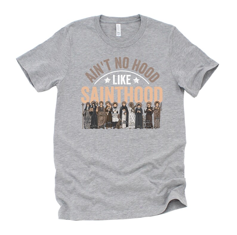 Pu&ograve; includere: T-shirt grigia con la scritta "Ain't No Hood Like Sainthood" in marrone chiaro e bianco. Il design include un'illustrazione di diversi santi in varie vesti.