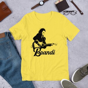 May include: A yellow t-shirt featuring a black and white graphic of a person playing a guitar, with the name "Brandi" in a stylized font below the image. The shirt is made of a soft material.