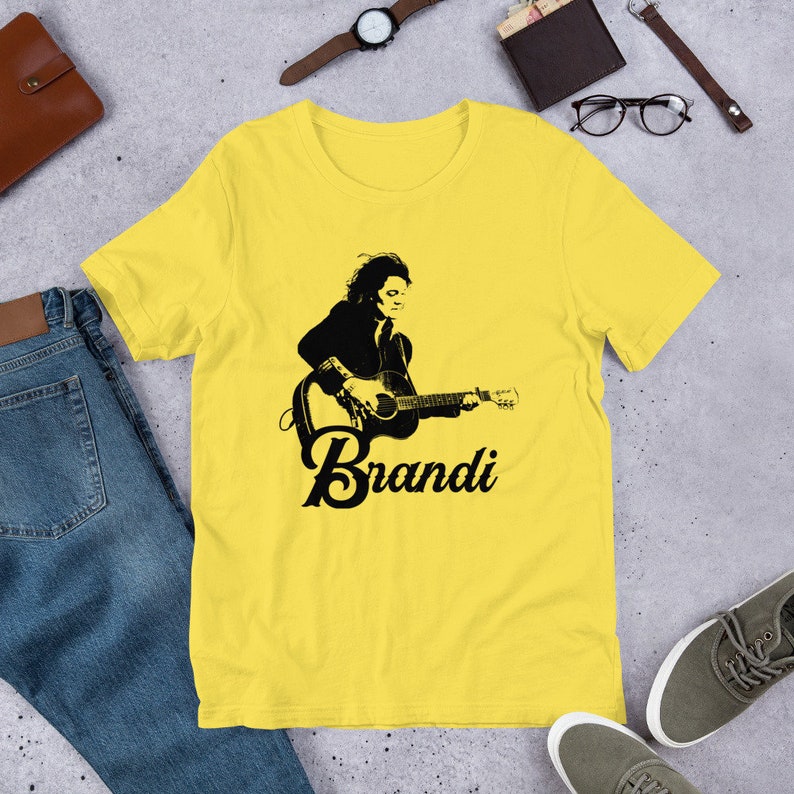 May include: A yellow t-shirt featuring a black and white graphic of a person playing a guitar, with the name "Brandi" in a stylized font below the image. The shirt is made of a soft material.