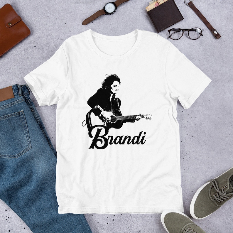 May include: White t-shirt featuring a black and white graphic of a person playing a guitar, with the name "Brandi" in a stylized font below the image. The shirt is made of a soft material.