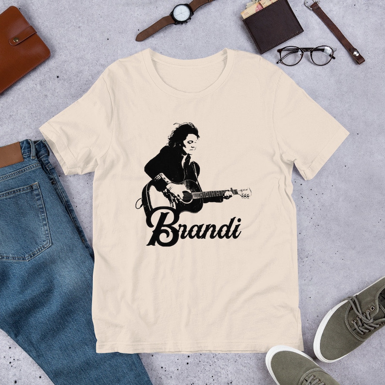 May include: Cream-colored t-shirt featuring a black and white graphic of a person playing a guitar, with the name "Brandi" in a stylized font below. The design is centered on the front of the shirt, creating a simple yet striking visual.