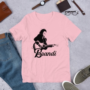 May include: Pink t-shirt featuring a black and white graphic of a person playing a guitar, with the name "Brandi" below. The shirt is made of soft material and is suitable for casual wear.