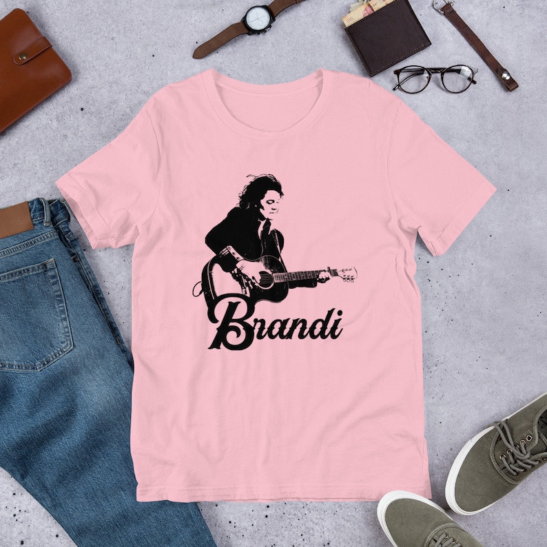 May include: Pink t-shirt featuring a black and white graphic of a person playing a guitar, with the name "Brandi" below. The shirt is made of soft material and is suitable for casual wear.