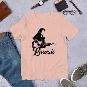 May include: A light pink t-shirt featuring a black and white graphic of a person playing a guitar, with the name "Brandi" below. The shirt is made of soft material and is suitable for casual wear.
