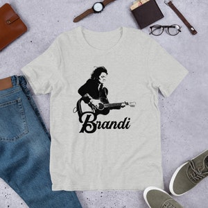 May include: A light gray t-shirt featuring a black and white graphic of a person playing a guitar, with the name "Brandi" printed below. The shirt is made of a soft material and is suitable for casual wear.