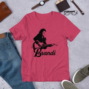 May include: A red t-shirt featuring a black graphic of a person playing a guitar, with the name "Brandi" in a script font below the image. The shirt is made of a soft material.