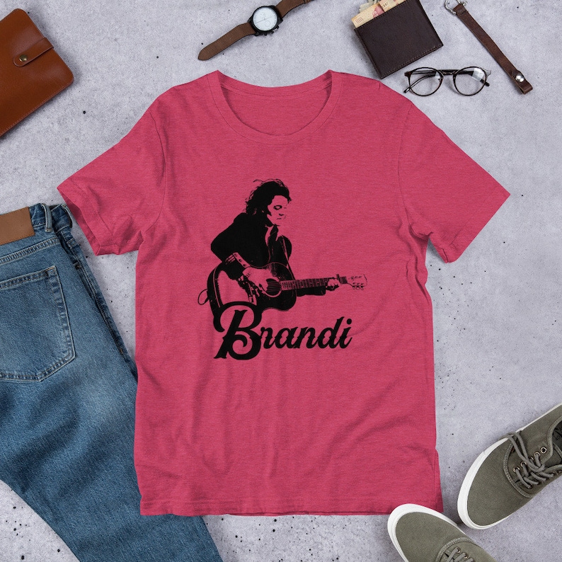 May include: A red t-shirt featuring a black graphic of a person playing a guitar, with the name "Brandi" in a script font below the image. The shirt is made of a soft material.