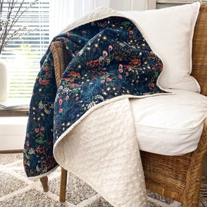 Peut inclure: Une couverture bleu marine et crème à motifs floraux est drapée sur une chaise en osier. La couverture présente un fond bleu foncé avec un motif floral coloré et un envers texturé crème. Un coussin blanc repose sur la chaise.