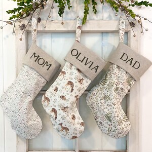 May include: Three Christmas stockings with beige tops and patterned bodies hang against a white background. The stockings are labelled "MOM", "OLIVIA", and "DAD". The patterns include a floral design, deer, and fern leaves.