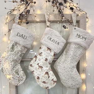 May include: Three Christmas stockings with white faux fur cuffs. One stocking has the word "DAD" embroidered, another "OLIVIA", and the third "MOM". The stockings have different patterns, including ferns and deer.