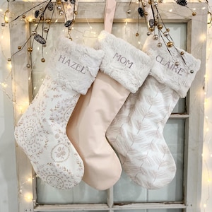May include: Three Christmas stockings with plush white faux fur cuffs. The left stocking features a floral pattern, the centre is solid light pink, and the right has a leaf print. Embroidered names read HAZEL, MOM, and CLAIRE respectively.