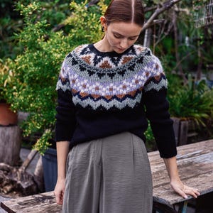Può includere: Un maglione nero con collo a fantasia nei toni del marrone, blu e bianco. Il maglione è abbinato a pantaloni verde oliva. L'immagine è scattata all'aperto, con vegetazione sullo sfondo.