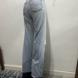 May include: Light blue denim jeans with a distressed knee detail. The jeans have a classic five-pocket design and a red Levi's tag on the back pocket. The jeans are paired with black boots.