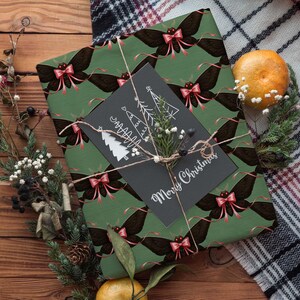 May include: A wrapped gift with green paper featuring a repeating pattern of black moths with pink bows. A black card with white Christmas tree illustrations and "Merry Christmas" text is tied with twine. The scene includes greenery and a tangerine.