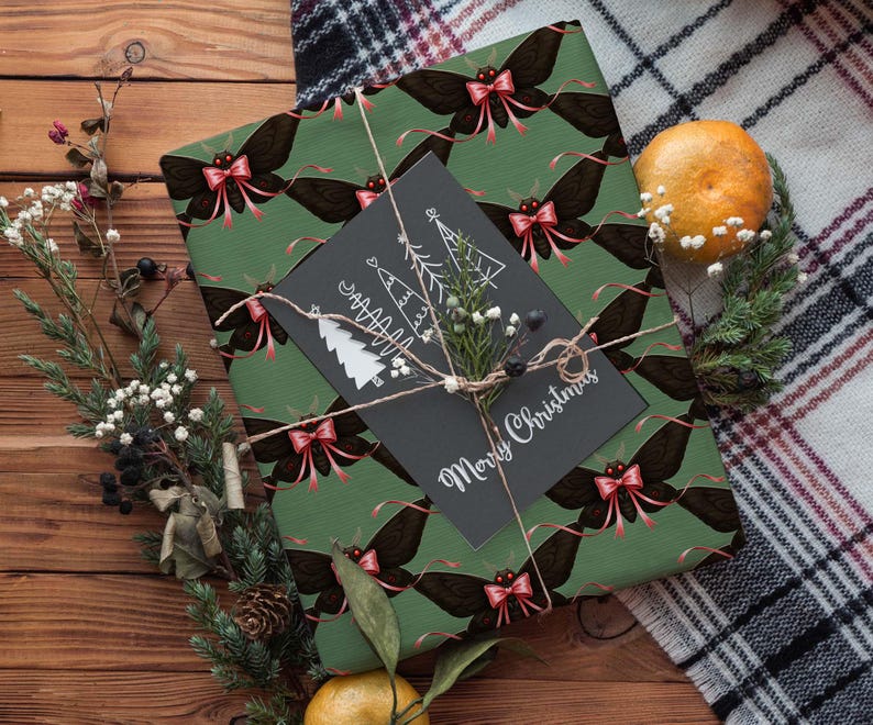 May include: A wrapped gift with green paper featuring a repeating pattern of black moths with pink bows. A black card with white Christmas tree illustrations and "Merry Christmas" text is tied with twine. The scene includes greenery and a tangerine.