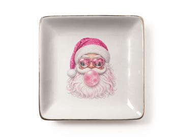Può includere: Un piattino quadrato in ceramica bianca con un bordo dorato metallizzato. Al centro è presente un'illustrazione grafica di Babbo Natale con un cappello rosa, occhiali a forma di cuore e una grande bolla di gomma da masticare rosa.